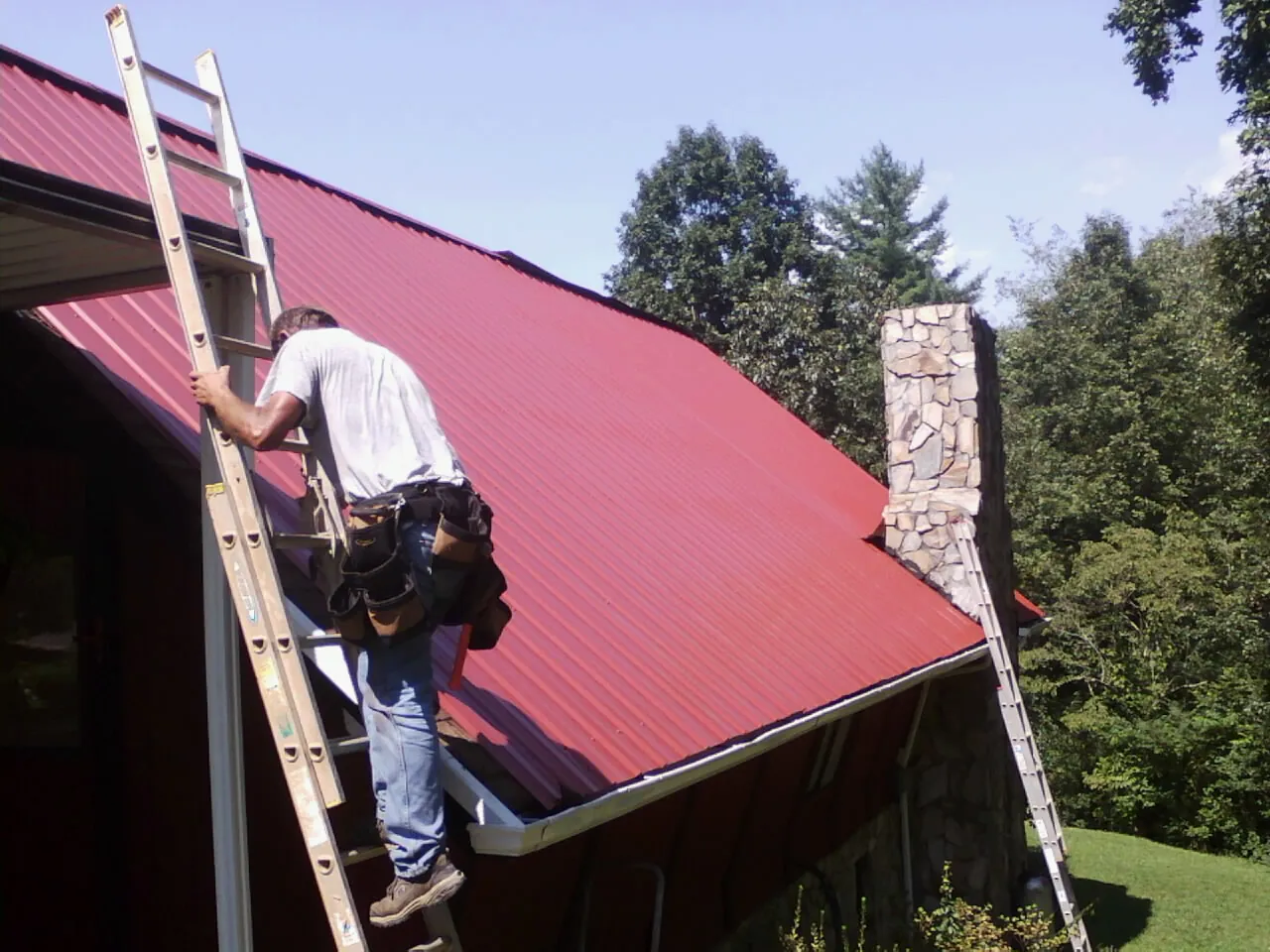 Roofer on a ladder beside a red metal roof and stone chimney — Soffit & Fascia Repair by Roofing Brothers in Keystone Heights